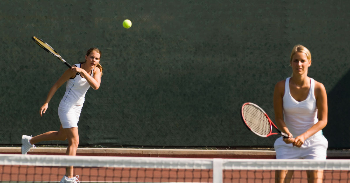 Two girls playing tennis doubles wearing white tennis outfits shown returning the ball