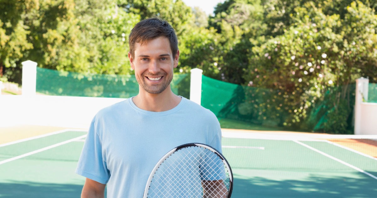 Young man with short brown hair wearing light blue t-shirt holding tennis racket in front
