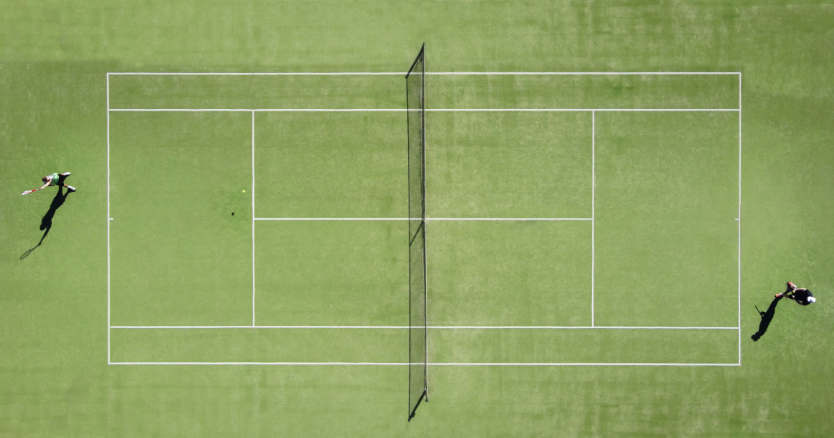 Overhead shot of green grass tennis court showing a player on either end of the baseline preparing to play a point