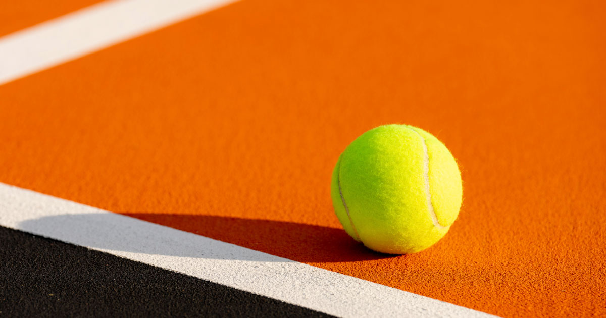 Up close photo of yellow tennis ball sitting on orange and black court with white lines
