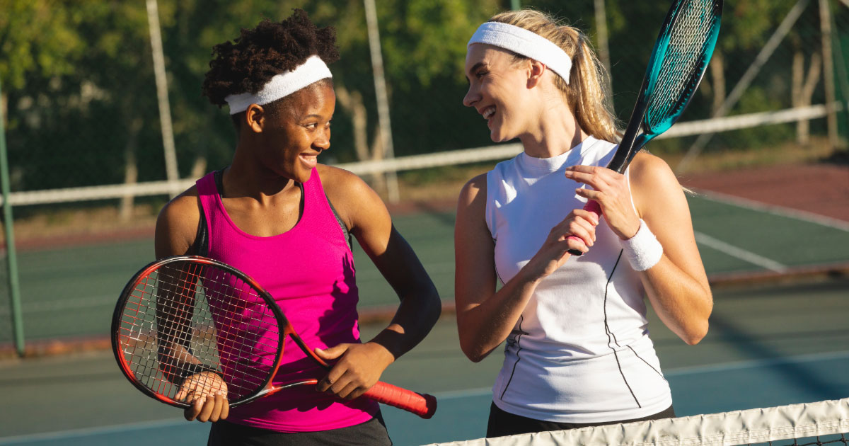 Two teen tennis girls wearing headbands and tank tops having a laugh holding their rackets at the net