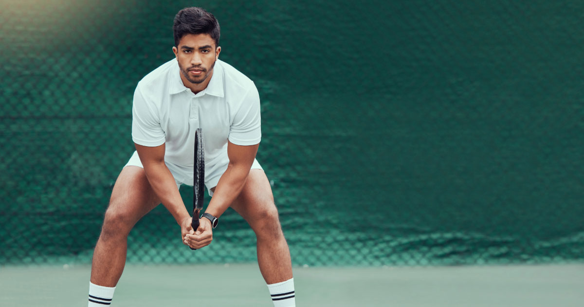 Young Spanish man wearing whtie shirt and shorts holding tennis racket with intense look on his face as he awaits his opponent's serve