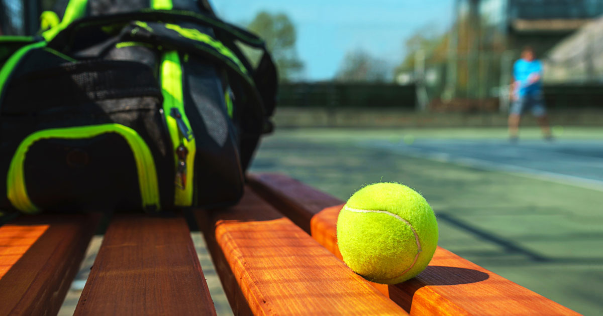 Close up photo of black and green tennis bag and ball on bench with blurred player in distance
